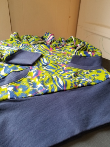 [Image;Colorful top taken at the angle of the waistband looking up on a flat lay. You can see the blue bands of the cuff and waistband and a cowl in the distance. The bands are blue and the rest of the top is a brightly colored animal print with lime green, blue, pink and grasy]