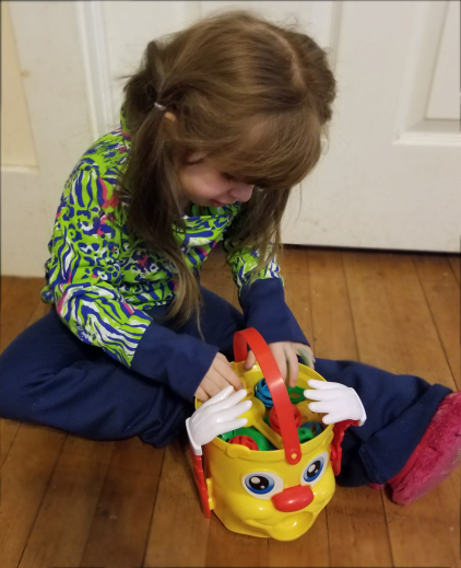 [Image: Baby O pulls balls out of her Mr. Bucket toy while sitting on the floor. One leg is tucked close to her body the other is straight out next to the Mr. Bucket]